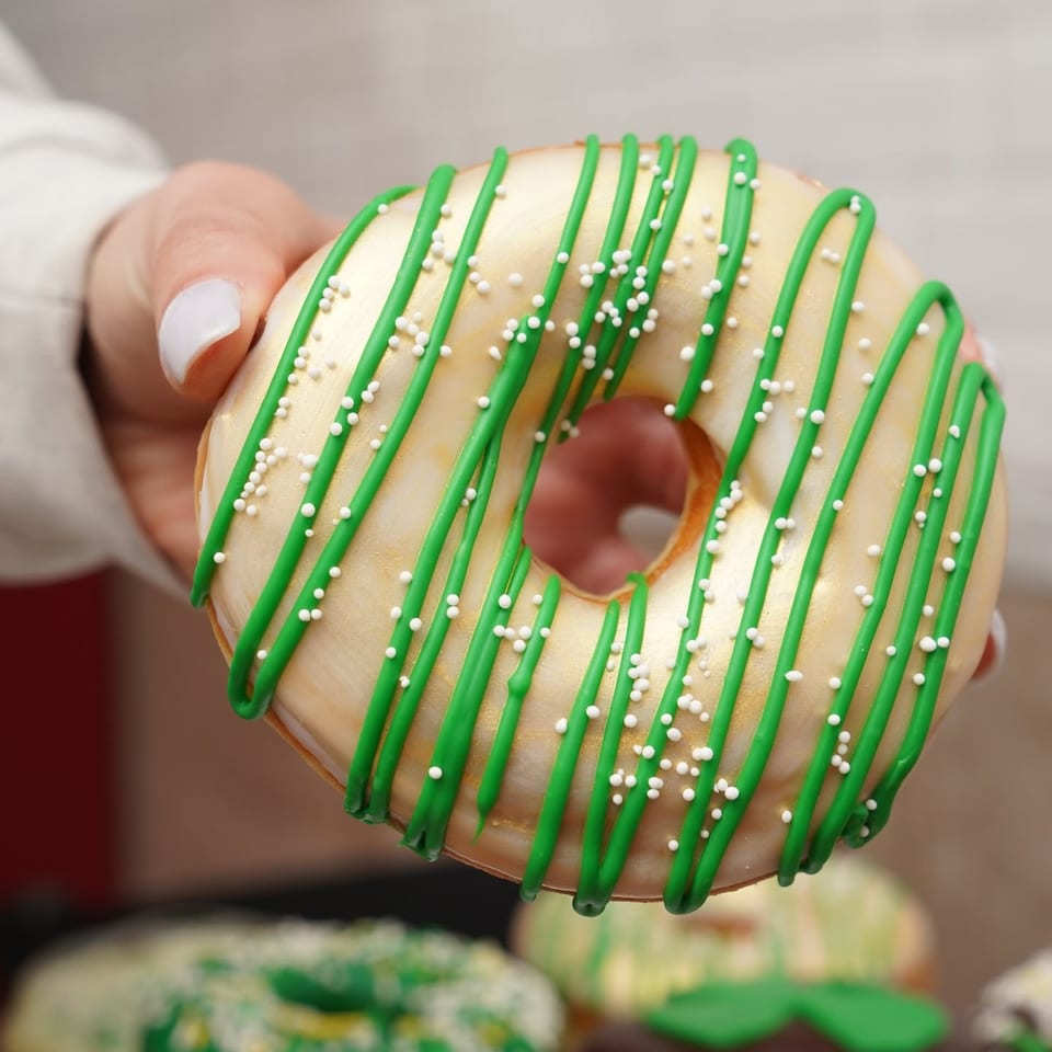 Glazed ring doughnut drizzled with green icing and covered in white sprinkles, held by a hand.
