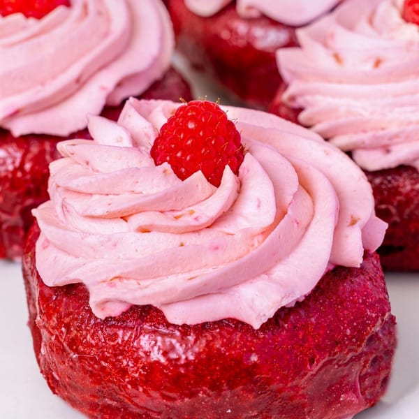 Close-up of a vibrant red donut topped with pink whipped cream and a fresh raspberry.