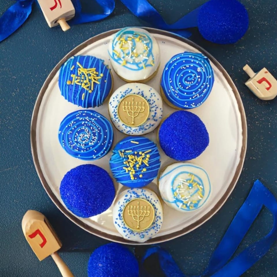 Blue and white Hanukkah cookies decorated with menorahs, swirls, sprinkles, and gold crumbs on a plate surrounded by dreidels and ribbon.
