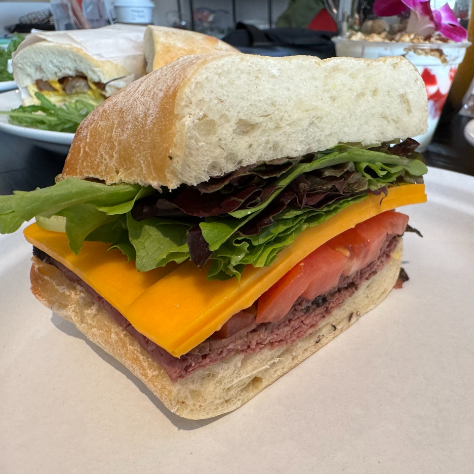 Deli sandwich on rustic bread layered with roast beef, cheddar cheese, mixed greens, and tomato on a restaurant table.