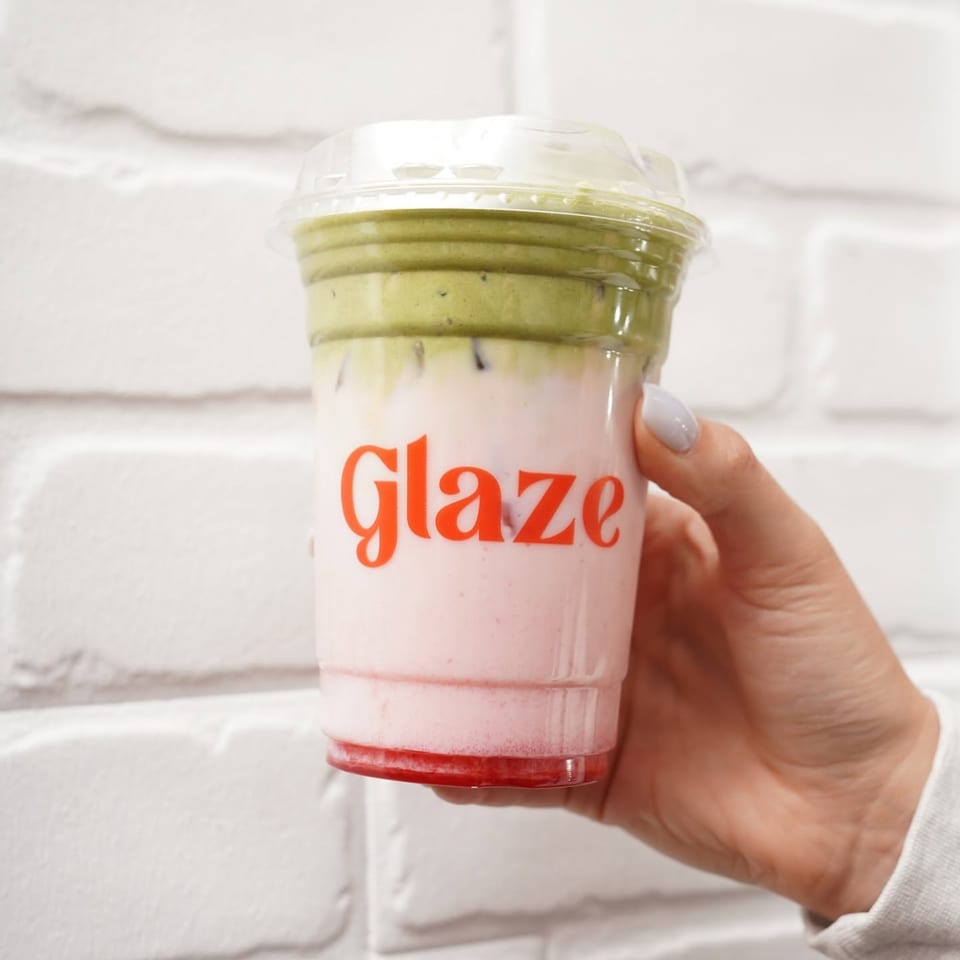 Hand holding a clear cup of layered pink and green smoothie labeled “glaze” against a white brick wall.