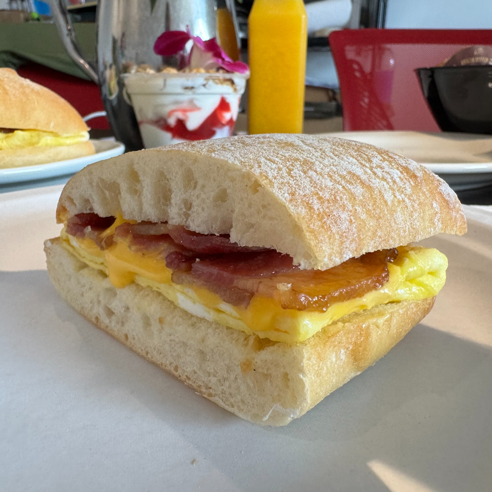 Breakfast sandwich with egg, melted cheese, bacon, and tomato on a flour-dusted ciabatta roll, served with dessert and juice.