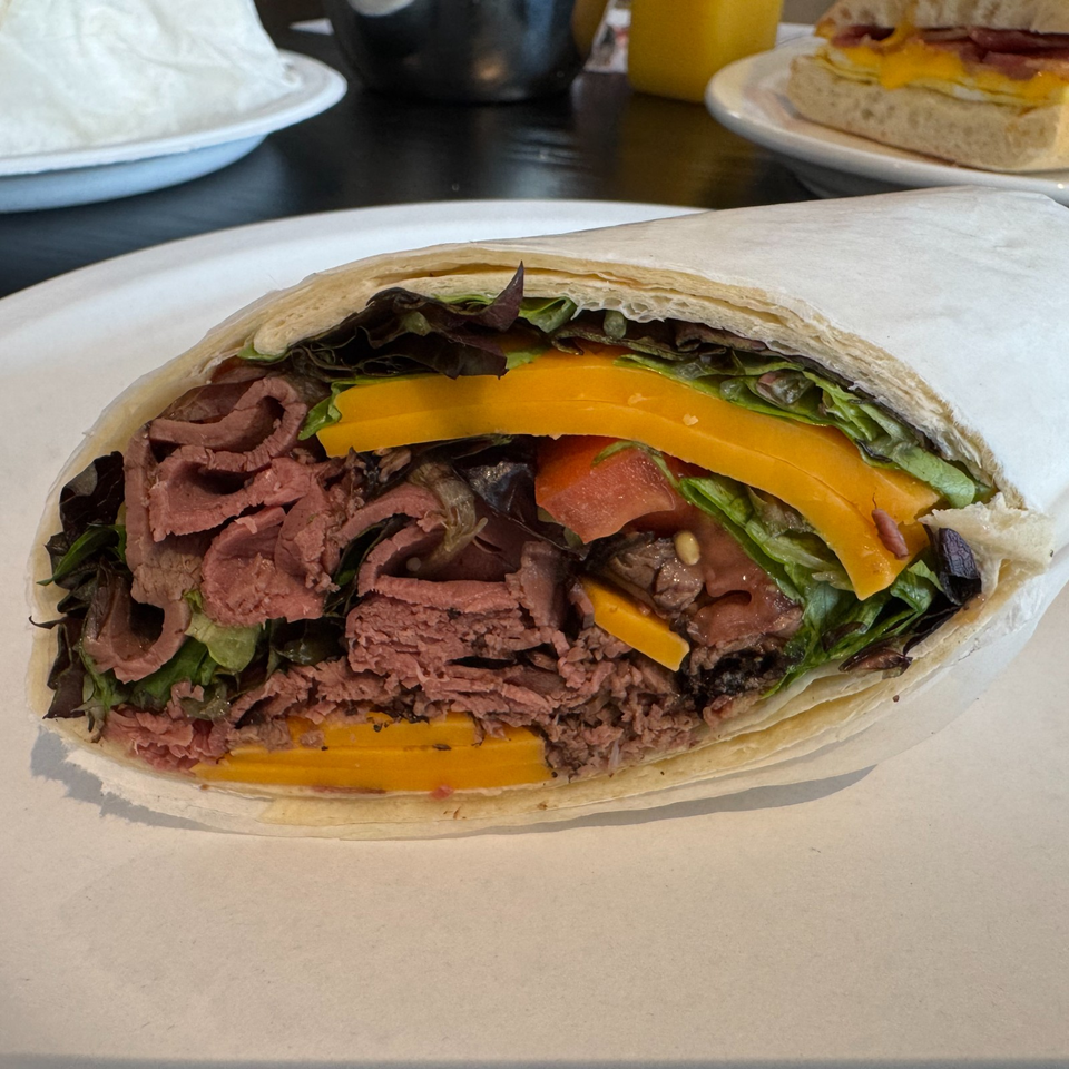Open flatbread sandwich filled with sliced roast beef, leafy greens, orange bell pepper, tomatoes, and mushrooms on a plate.