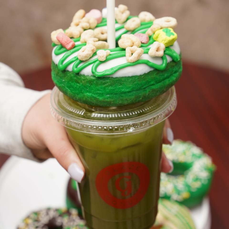 Hand holding a Glaze Donuts matcha drink topped with a green and white frosted donut.