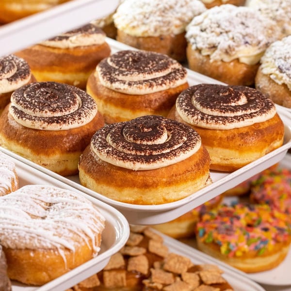 Trays of freshly baked donuts, some topped with chocolate powder swirls, others with powdered sugar or colorful sprinkles.