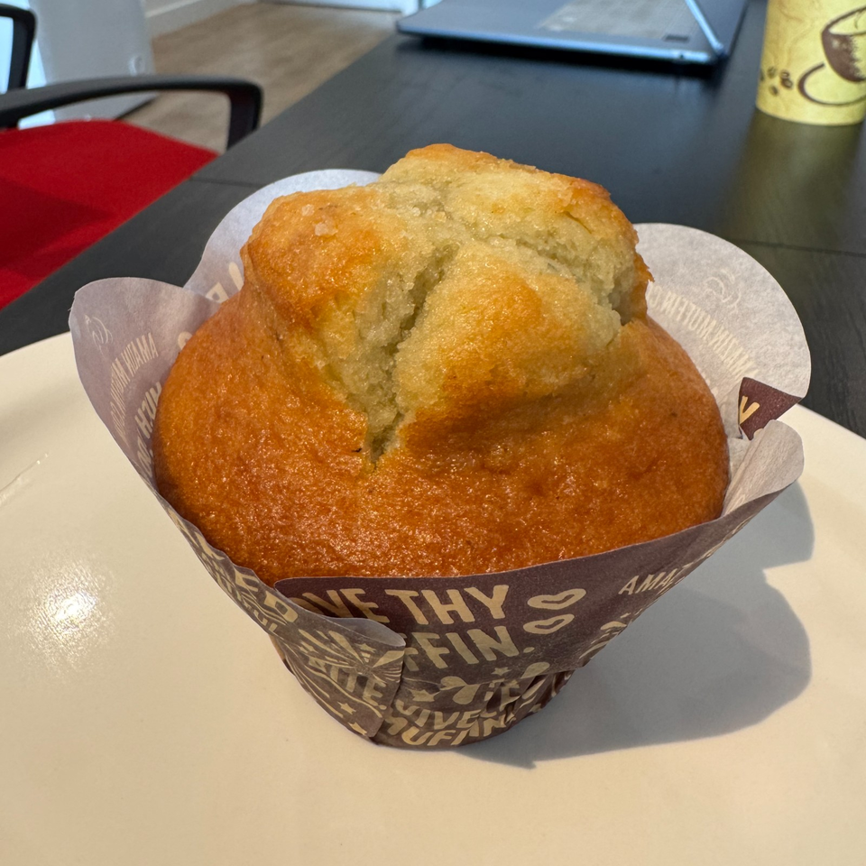 Golden-brown muffin with a cracked, pale center in a branded paper wrapper on a café table.