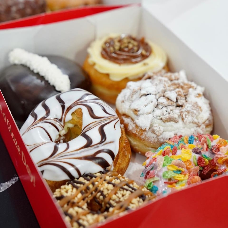 A red and white box filled with a variety of gourmet donuts, including frosted, powdered, and cereal-topped.