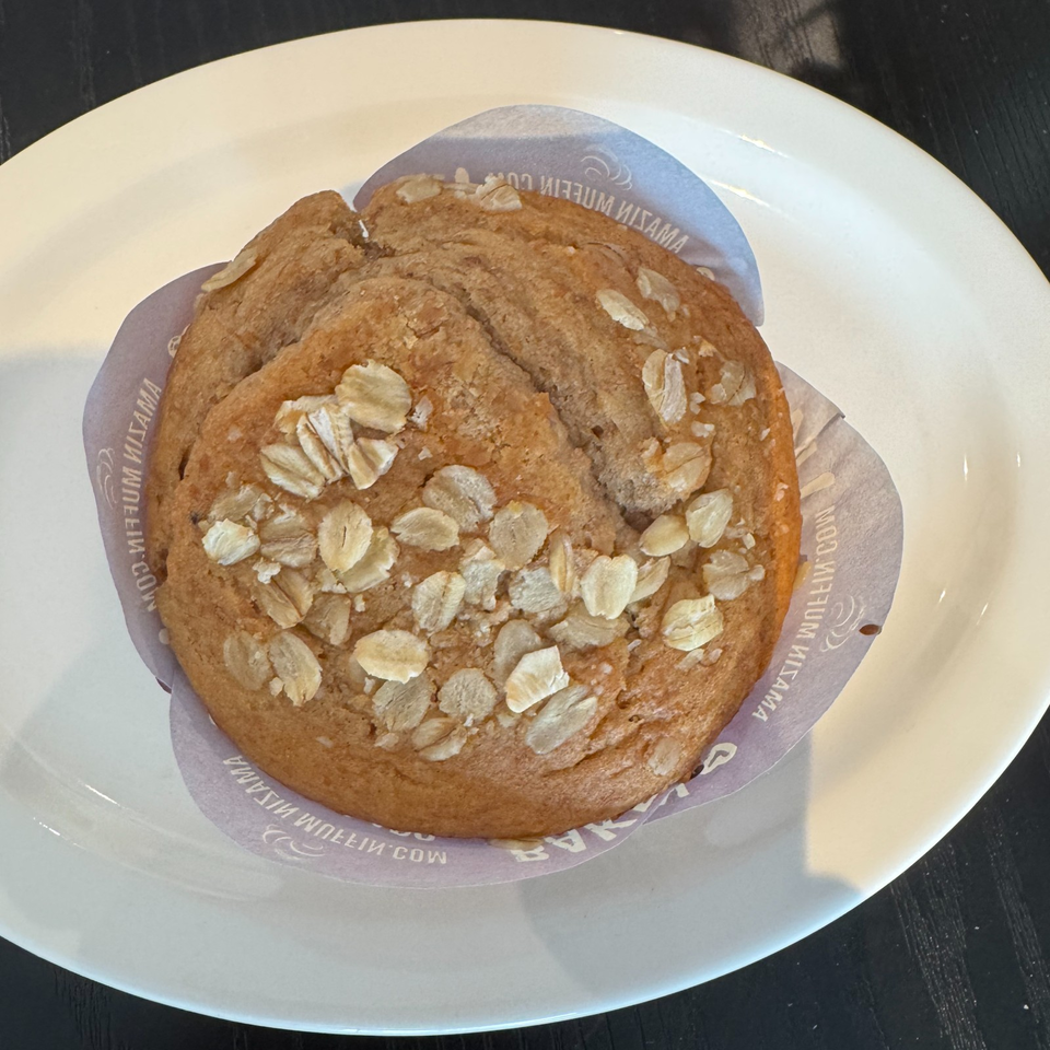 Golden-brown oat muffin topped with rolled oats, served in a branded paper liner on a white plate.