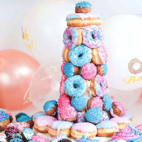 A tall tower of pink and blue frosted donuts and donut holes, with balloons in the background.