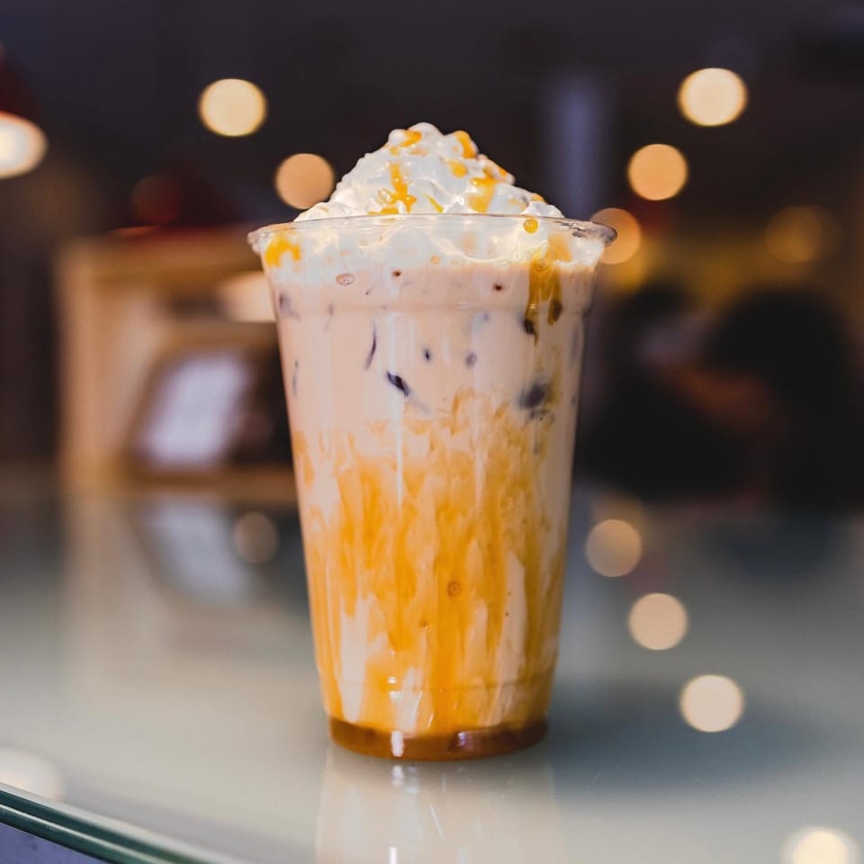 Caramel-drizzled milkshake topped with whipped cream in a glass, set against a warmly lit restaurant background.