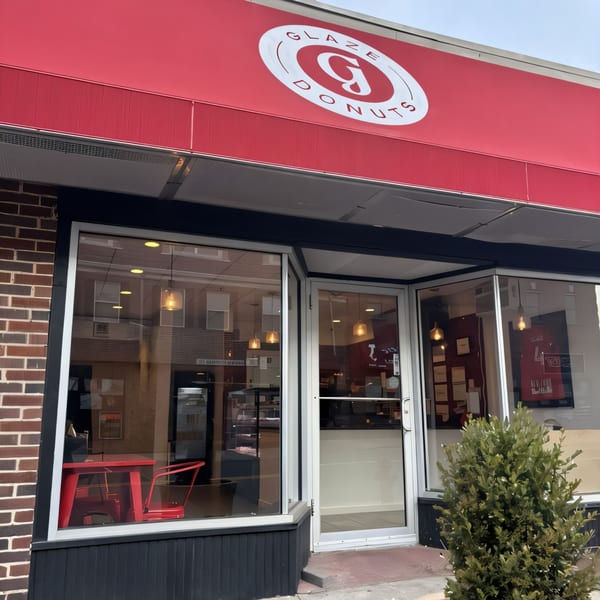 Exterior of Glaze Donuts, a brick building with a red awning and large windows showcasing red tables.