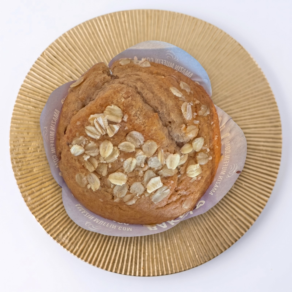 Oat-topped golden muffin in a branded paper liner on a ridged gold plate.