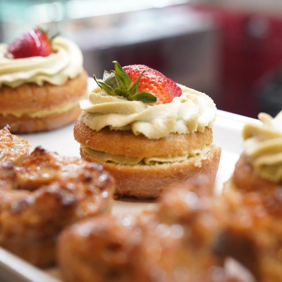 Close-up of a cream-topped pastry garnished with a fresh strawberry, surrounded by assorted pastries on a serving tray.