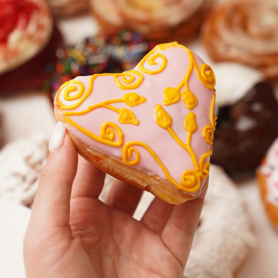 Hand holding a pink iced cookie decorated with intricate golden-orange swirls and floral patterns.